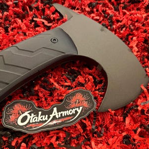 Puede incluir: Un hacha táctica de color gris oscuro mate con hoja curva y mango negro con patrón de chevrón. Una pegatina negra con ilustraciones de lirios araña rojos y el texto blanco Otaku Armory se encuentra cerca de la cabeza del hacha.