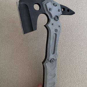 May include: A tactical-style hatchet featuring a matte black blade with a sharpened edge, a circular cutout, and a pointed rear spike. The handle and head assembly are constructed from layered gray and black components secured with visible hex bolts.