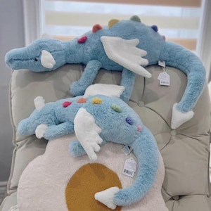 May include: Two plush blue dragon toys with white wings and colourful accents. The dragons have long tails and are lying on a neutral-coloured chair. The toys have small tags attached.