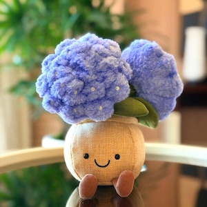 May include: A plush toy shaped like a smiling flower pot with blue hydrangea blooms. The pot is tan with a stitched face and small brown feet. The blue flowers have white accents and green leaves.