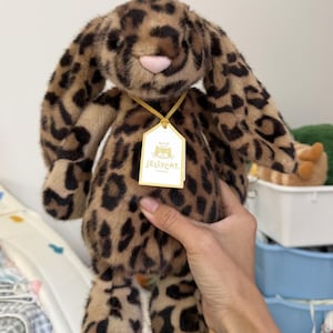 Può includere: Un coniglietto di peluche con stampa leopardata, lunghe orecchie flosce e un naso rosa. Il peluche ha un'etichetta con la scritta "Jellycat London". Il coniglietto è morbido e coccoloso.