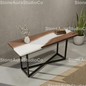 Puede incluir: Una mesa de centro rectangular con una tapa de nogal de borde vivo y un centro de resina epoxi blanca. La mesa tiene una base de metal negro y está decorada con un jarrón blanco y libros. La mesa mide aproximadamente 152 cm de largo.