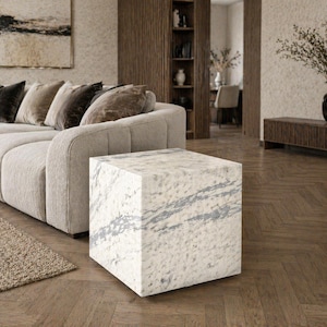 May include: A square marble side table with gray veining. The table is in a modern living room setting, next to a beige sofa with several throw pillows. The room has a neutral color palette with wooden accents.