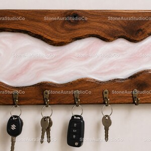 Peut inclure: Porte-clés en bois avec une rivière de résine blanche et rose. Le porte-clés comporte cinq crochets en bronze et peut contenir plusieurs clés. Le bois présente un motif de grain naturel. Le texte "StoneAuraStudioCo" est visible.