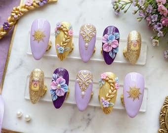 Op de afbeelding: Een set op maat gemaakte, lange, ovaalvormige press-on nagels. De nagels hebben een lichtpaarse basis met gouden accenten, bloemmotieven en een zonnesymbool. Sommige nagels hebben een glittergouden afwerking, andere 3D-bloemversieringen.