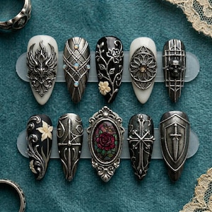 May include: A collection of ten gothic-style press-on nails. Designs include ornate silver embellishments, floral patterns, a skull, and a stained-glass rose. The nails are in various shapes and colors, including black, white, and silver.