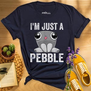 May include: A navy blue t-shirt featuring a graphic of a grey, round, cartoon-style pebble character with large eyes and a small tongue sticking out. The text "I'M JUST A PEBBLE" is printed in white capital letters above and below the character.