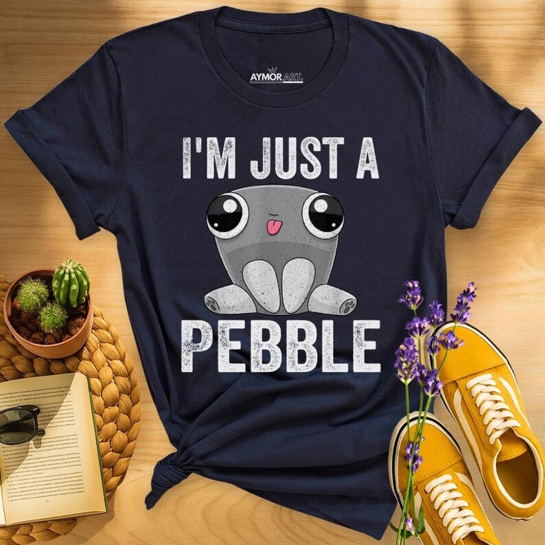 May include: A navy blue t-shirt featuring a graphic of a grey, round, cartoon-style pebble character with large eyes and a small tongue sticking out. The text "I'M JUST A PEBBLE" is printed in white capital letters above and below the character.