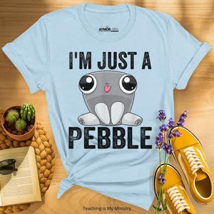 May include: A light blue short-sleeve t-shirt featuring a graphic of a grey cartoon creature with large eyes and a small tongue sticking out. The text above the creature reads I'M JUST A and the text below reads PEBBLE in black block letters.