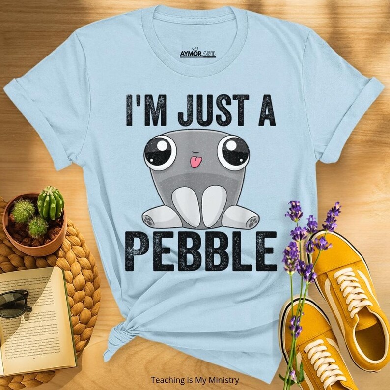 May include: A light blue short-sleeve t-shirt featuring a graphic of a grey cartoon creature with large eyes and a small tongue sticking out. The text above the creature reads I'M JUST A and the text below reads PEBBLE in black block letters.