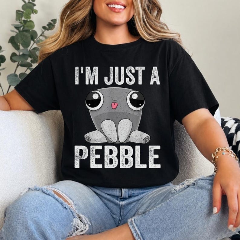 May include: A black short-sleeved t-shirt featuring a graphic of a grey, round, cartoon-style creature with large eyes and a small tongue sticking out. The text "I'M JUST A PEBBLE" is printed in white, distressed capital letters above and below.