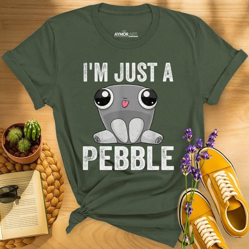 May include: An olive green short-sleeved t-shirt featuring a graphic of a grey cartoon creature with large eyes and a small tongue sticking out. The text "I'M JUST A PEBBLE" is printed in white above and below the creature. The brand "AYMOR ART" is visible.