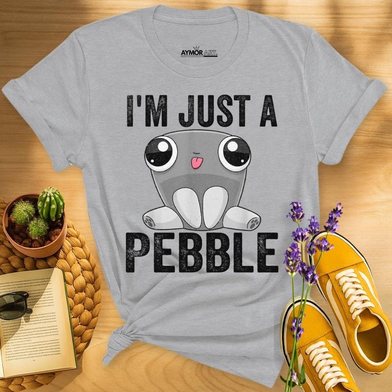 May include: A light gray short-sleeve t-shirt featuring a graphic of a cartoon gray pebble character with large eyes and a small tongue sticking out. The text "I'M JUST A PEBBLE" is printed in black above and below the character, with "AYMOR ART" at the top.