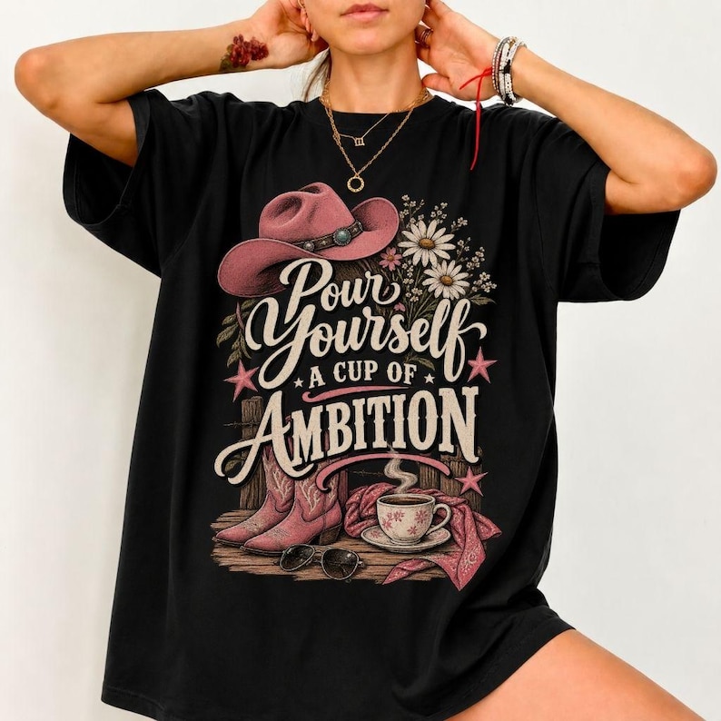 May include: A black t-shirt featuring a graphic print of a pink cowboy hat, a pair of pink cowboy boots, a coffee cup on a saucer, and white daisies. The text reads, "Pour Yourself A Cup Of Ambition." The design includes small pink stars.