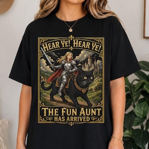 Puede incluir: Una camiseta negra con un estampado gráfico de un caballero con armadura plateada montado sobre un gato negro grande. El diseño incluye un castillo de fondo, una capa roja y el texto: HEAR YE! HEAR YE! THE FUN AUNT HAS ARRIVED, en un marco dorado.