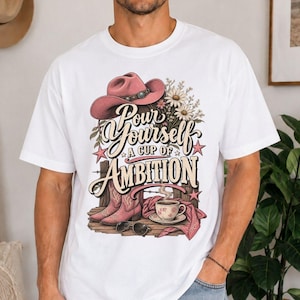 May include: A white t-shirt featuring a graphic print of a pink cowboy hat, a bouquet of daisies, a pair of pink cowboy boots, a coffee cup on a saucer, and sunglasses. The text reads, "Pour Yourself A CUP OF AMBITION" in stylised lettering.