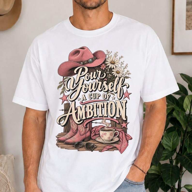 May include: A white t-shirt featuring a graphic print of a pink cowboy hat, a bouquet of daisies, a pair of pink cowboy boots, a coffee cup on a saucer, and sunglasses. The text reads, "Pour Yourself A CUP OF AMBITION" in stylised lettering.