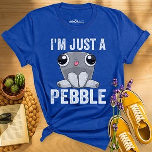 May include: A royal blue short-sleeve t-shirt featuring a graphic of a gray, round, cartoon-style creature with large eyes and a small tongue sticking out. The text "I'M JUST A PEBBLE" is printed in white, distressed capital letters above and below.