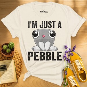 May include: A cream-colored short-sleeve t-shirt featuring a graphic of a gray cartoon pebble character with large eyes and a small tongue sticking out. The text "I'M JUST A PEBBLE" is printed in black block letters above and below the character.