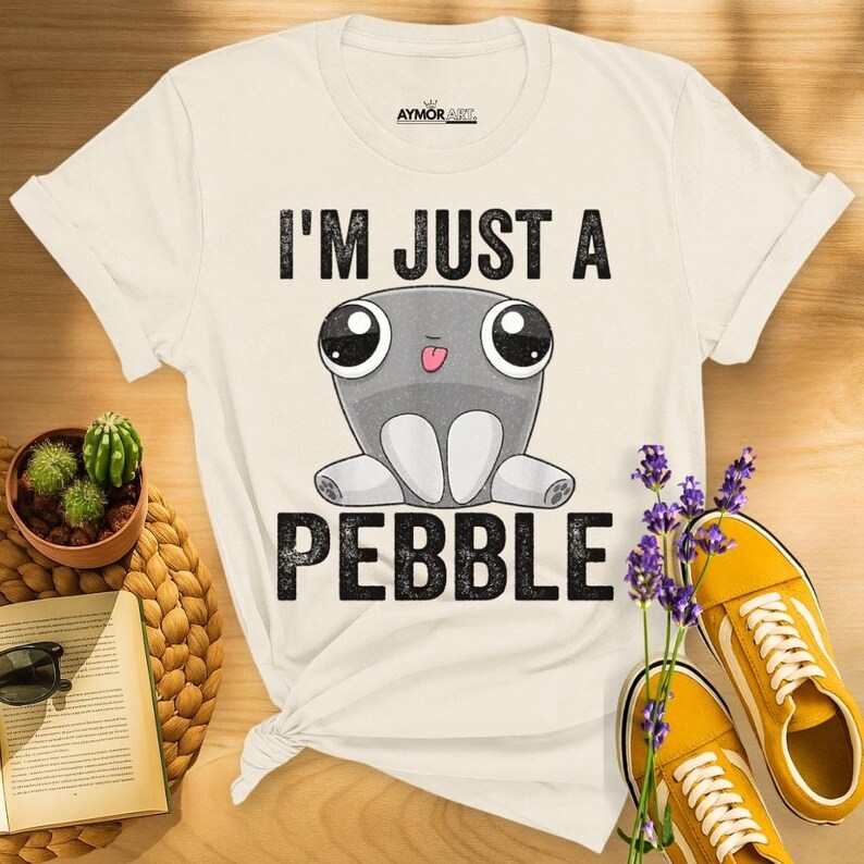 May include: A cream-colored short-sleeve t-shirt featuring a graphic of a gray cartoon pebble character with large eyes and a small tongue sticking out. The text "I'M JUST A PEBBLE" is printed in black block letters above and below the character.