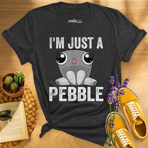 May include: A dark gray short-sleeve t-shirt featuring a graphic of a cartoon pebble character with large eyes and a small tongue sticking out. The text "I'M JUST A PEBBLE" is printed in white above and below the character. The shirt has a crew neck.