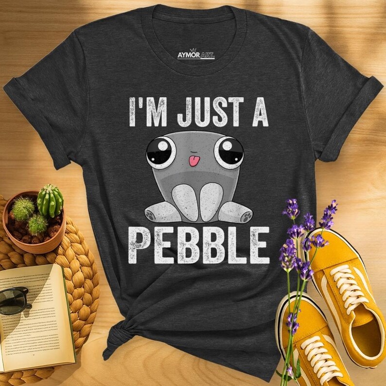 May include: A dark gray short-sleeve t-shirt featuring a graphic of a cartoon pebble character with large eyes and a small tongue sticking out. The text "I'M JUST A PEBBLE" is printed in white above and below the character. The shirt has a crew neck.