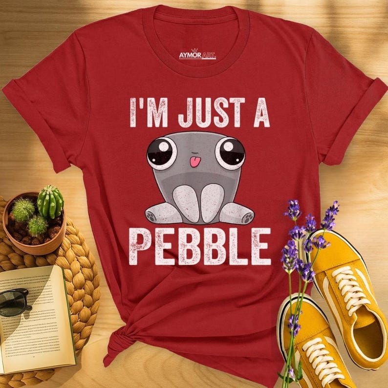 May include: A red short-sleeved t-shirt featuring a graphic of a grey, round creature with large eyes and a small tongue sticking out. The text "I'M JUST A PEBBLE" is printed in white above and below the creature. The brand name AYMOR ART is visible.