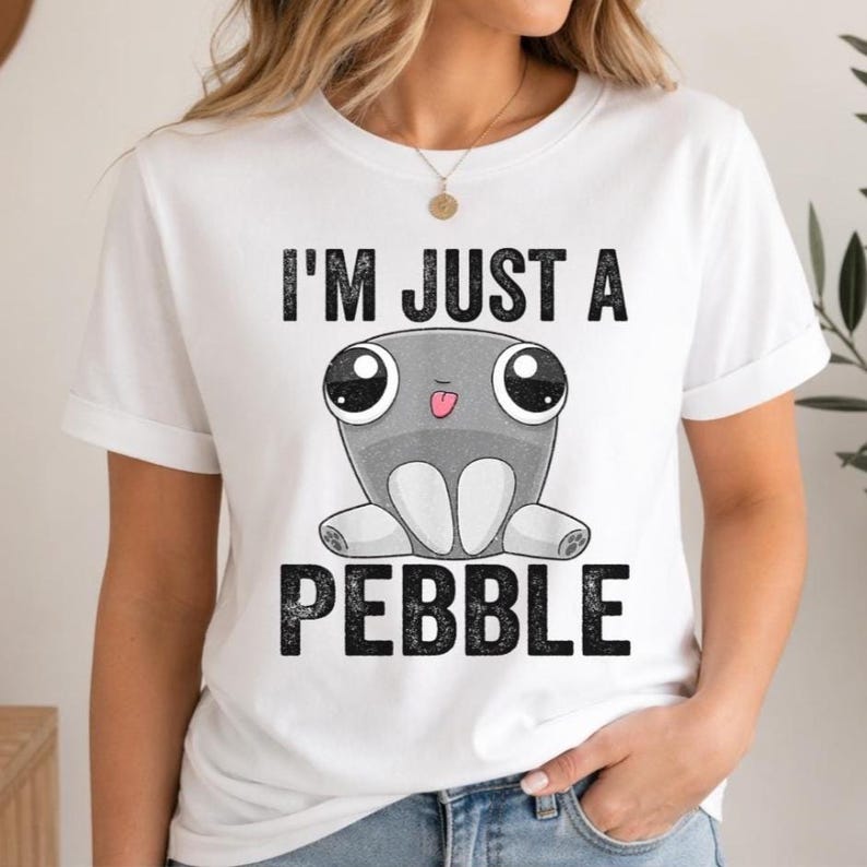 May include: A white short-sleeved t-shirt featuring a graphic of a grey, round, cartoon-style pebble character with large eyes and a small pink tongue sticking out. The text "I'M JUST A PEBBLE" is printed in black, distressed capital letters.