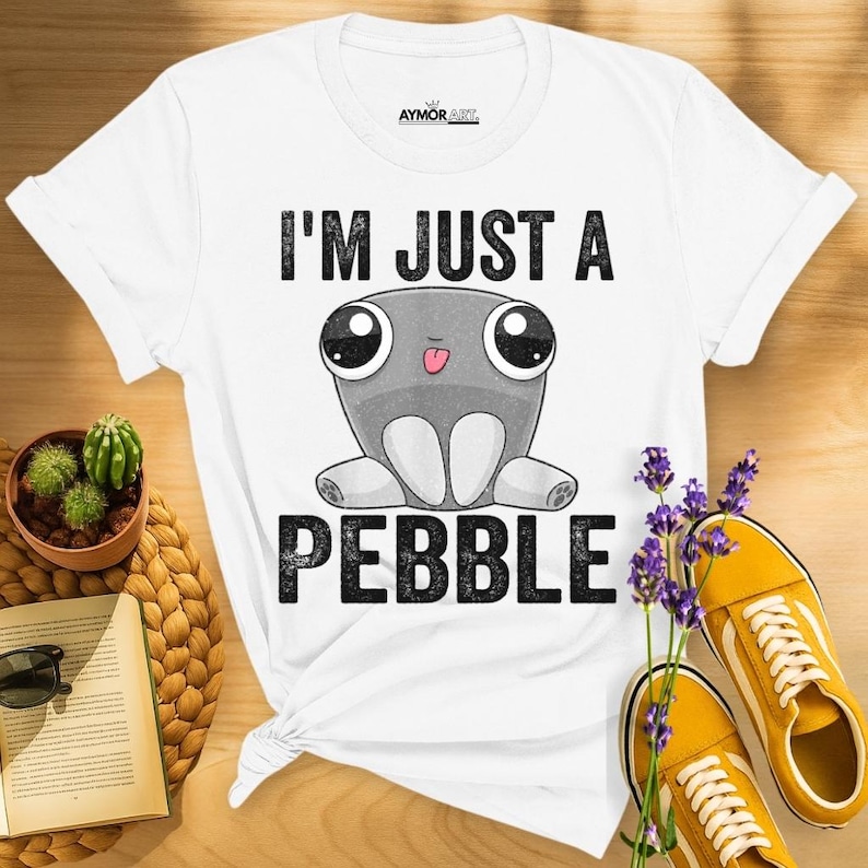 May include: A white short-sleeve t-shirt featuring a graphic of a gray cartoon pebble character with large eyes and a small tongue sticking out. The text "I'M JUST A PEBBLE" is printed in black capital letters above and below the character.