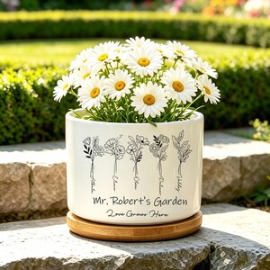 Könnte beinhalten: Ein weißer Keramik-Blumentopf mit einer Bambus-Untersetzer, gefüllt mit Gänseblümchen. Der Topf hat ein Blumendesign und den Text "Mr. Robert's Garden Love Grows Here". Der Topf steht auf einer Steinoberfläche in einem Garten.