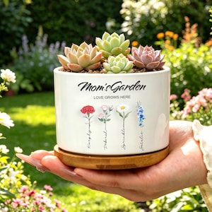Op de afbeelding: Een witte keramische plantenbak met de tekst "Mom's Garden" en "Love Grows Here" bevat verschillende vetplanten. De plantenbak heeft bloemenillustraties en staat op een houten basis. De achtergrond is een weelderige tuin.