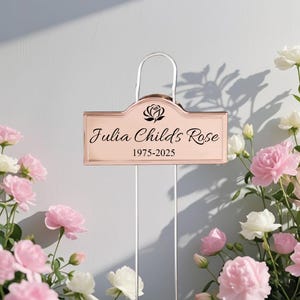 May include: A rose gold metal garden plant marker with a curved top, featuring a black engraved rose icon, the text "Julia Childs Rose", and the dates "1975-2025". The rectangular plaque is attached to two thin silver metal stakes.