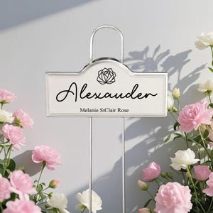 Puede incluir: Un letrero de jardín rectangular de metal plateado con un asa de alambre y dos estacas delgadas. El letrero muestra el nombre Alexander en letra cursiva negra, un icono de rosa geométrica y el texto Melanie StClair Rose debajo.