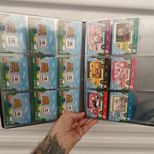 May include: A black card binder filled with trading cards. The cards feature colorful illustrations and text, with a variety of designs. The binder is held open, displaying the cards in clear plastic sleeves.