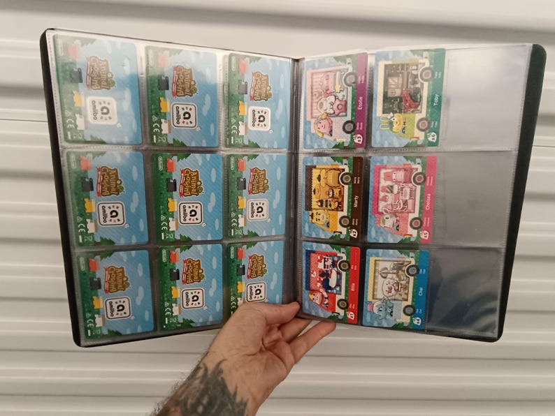 May include: A black card binder filled with trading cards. The cards feature colorful illustrations and text, with a variety of designs. The binder is held open, displaying the cards in clear plastic sleeves.