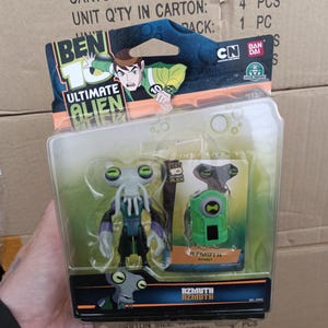 Puede incluir: Una figura de acci&oacute;n de Azmuth de Ben 10 Ultimate Alien en un paquete de pl&aacute;stico transparente. La figura es gris, morada y negra. El paquete tambi&eacute;n incluye un dispositivo verde. El embalaje presenta el logotipo de Ben 10 y un personaje de dibujos animados.