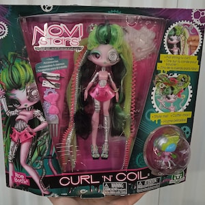 May include: A Novi Stars Curl 'N' Coil doll in a clear plastic package. The doll features green and black hair, a pink skirt, and silver metallic details. The packaging includes hair accessories and instructions.