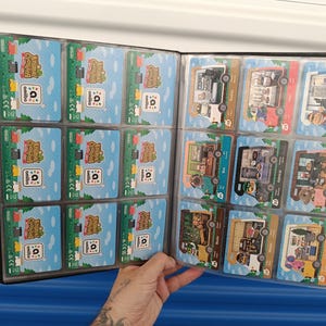 May include: A black card binder filled with Animal Crossing Amiibo cards. The cards feature colorful illustrations of characters and scenes from the game. The binder is open, displaying multiple cards in clear plastic sleeves.