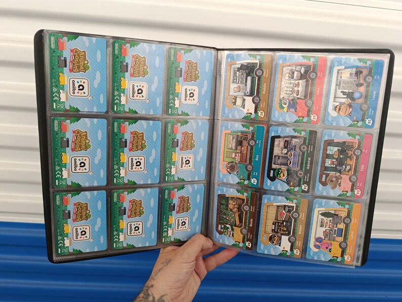 May include: A black card binder filled with Animal Crossing Amiibo cards. The cards feature colorful illustrations of characters and scenes from the game. The binder is open, displaying multiple cards in clear plastic sleeves.