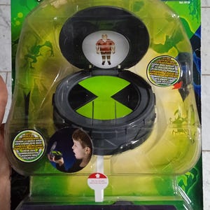 Könnte beinhalten: Ben 10 Alien Force Plumber's Communicator Spielzeug in einer transparenten Kunststoffverpackung. Das Spielzeug hat ein grünes und schwarzes Design mit einem kreisförmigen Zifferblatt, einem grünen X-Symbol und einem Charakterbild.