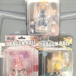 Puede incluir: Tres figuras de acci&oacute;n de Dragon Ball Z en embalaje de pl&aacute;stico transparente. Las figuras representan personajes con pelo amarillo y ropa azul, piel rosa y pantalones blancos, y ropa azul y naranja. Cada figura est&aacute; en una funda de pl&aacute;stico.