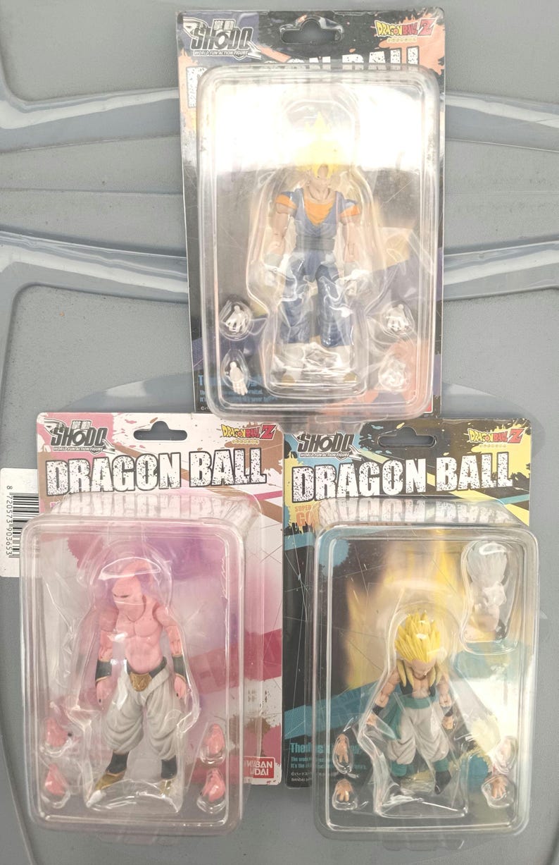 Puede incluir: Tres figuras de acci&oacute;n de Dragon Ball Z en embalaje de pl&aacute;stico transparente. Las figuras representan personajes con pelo amarillo y ropa azul, piel rosa y pantalones blancos, y ropa azul y naranja. Cada figura est&aacute; en una funda de pl&aacute;stico.