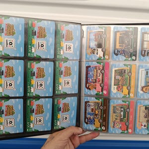 May include: A black card binder filled with Animal Crossing amiibo cards. The cards feature colorful illustrations of characters and scenes from the game. The binder is open, displaying multiple cards in clear plastic sleeves.