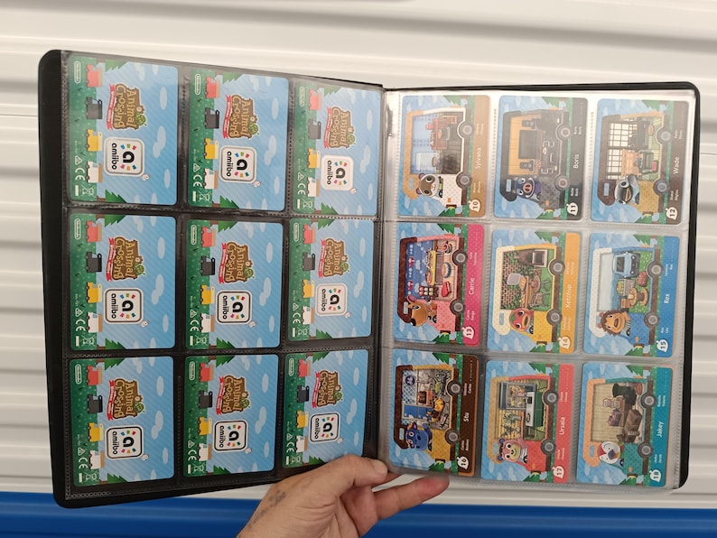 May include: A black card binder filled with Animal Crossing amiibo cards. The cards feature colorful illustrations of characters and scenes from the game. The binder is open, displaying multiple cards in clear plastic sleeves.
