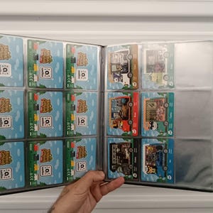 May include: A black trading card binder filled with colorful Animal Crossing amiibo cards. The cards feature various characters and scenes from the game, with a blue and green background. The binder is open, displaying multiple cards in clear plastic sleeves.