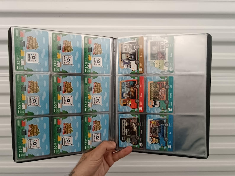 May include: A black trading card binder filled with colorful Animal Crossing amiibo cards. The cards feature various characters and scenes from the game, with a blue and green background. The binder is open, displaying multiple cards in clear plastic sleeves.