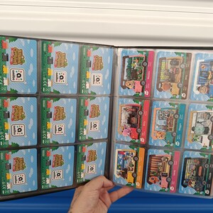 May include: A black card binder filled with Animal Crossing Amiibo cards. The cards feature colorful illustrations of characters and scenes from the game. The binder is open, displaying multiple pages of cards in clear plastic sleeves.