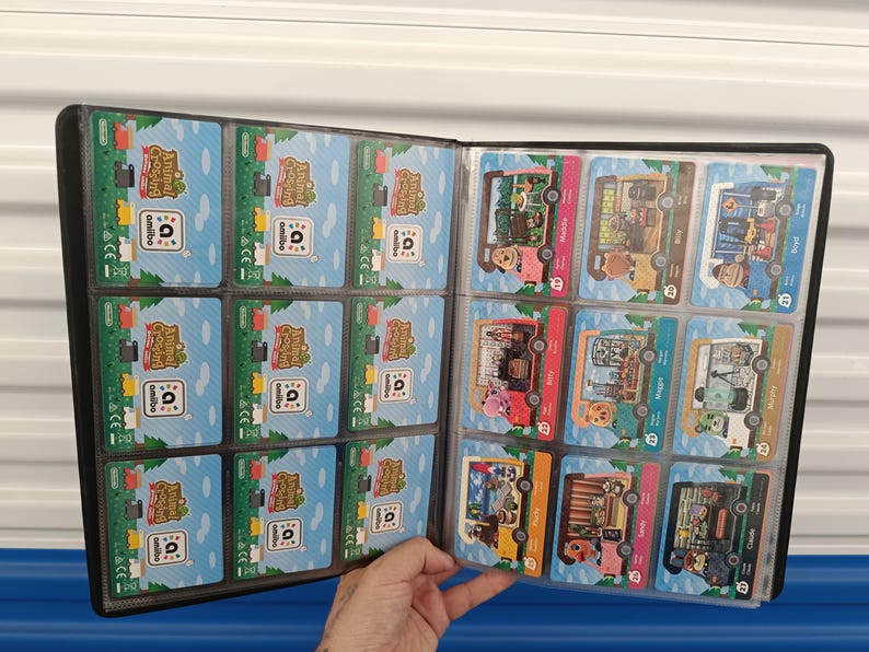 May include: A black card binder filled with Animal Crossing Amiibo cards. The cards feature colorful illustrations of characters and scenes from the game. The binder is open, displaying multiple pages of cards in clear plastic sleeves.