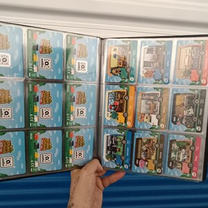 May include: A black card binder filled with Animal Crossing Amiibo cards. The cards feature colorful illustrations of characters and buildings on a blue and green background. The binder is open, displaying multiple cards in clear plastic sleeves.
