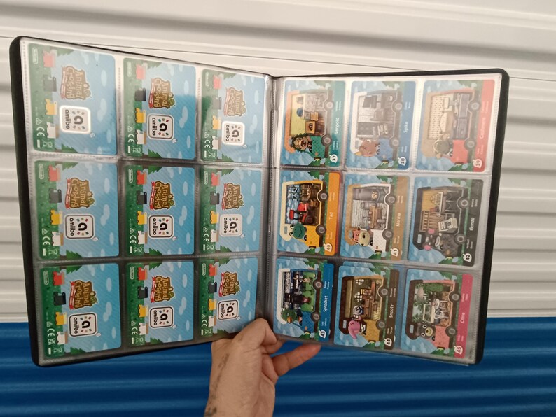 May include: A black card binder filled with Animal Crossing Amiibo cards. The cards feature colorful illustrations of characters and buildings on a blue and green background. The binder is open, displaying multiple cards in clear plastic sleeves.